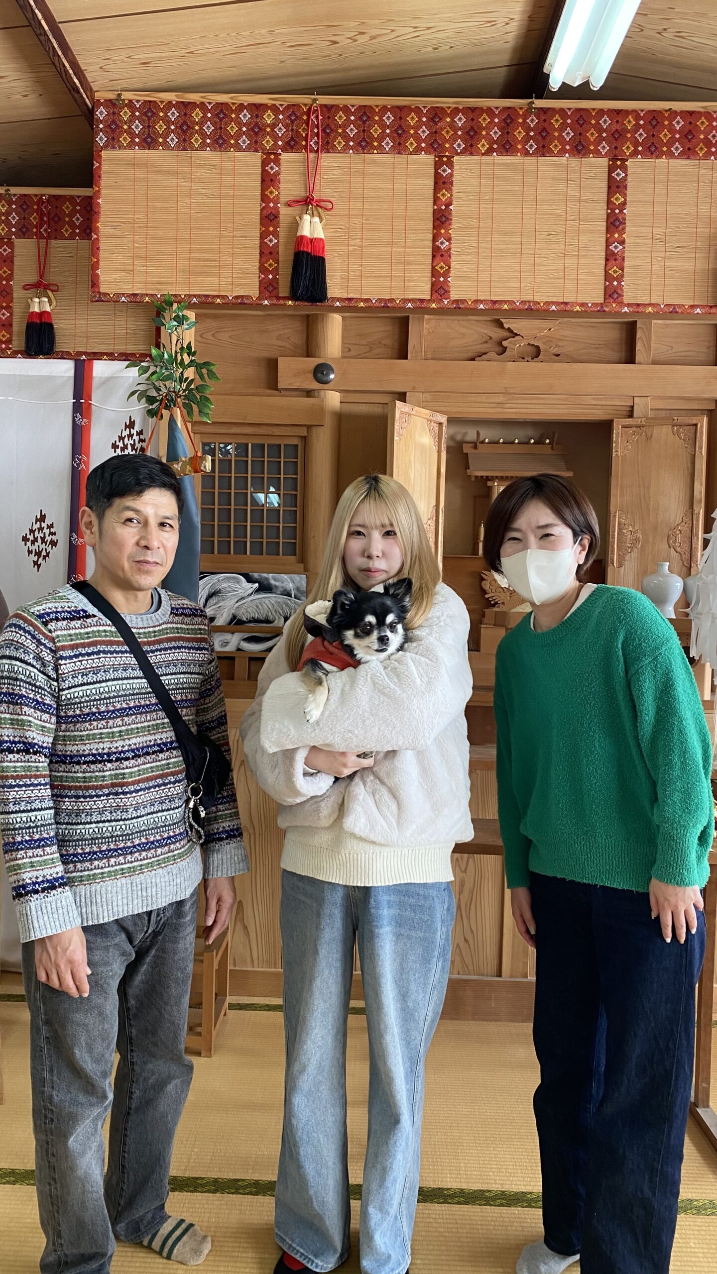 畳の部屋で左にセーターとジーンズを着た男性、中央に白いもこもこジャケットとジーンズを着て小型犬を抱える女性、右に緑のセーターとマスクを着けた女性が立っている。背後には和風の木製の飾りと伝統的な装飾がある。