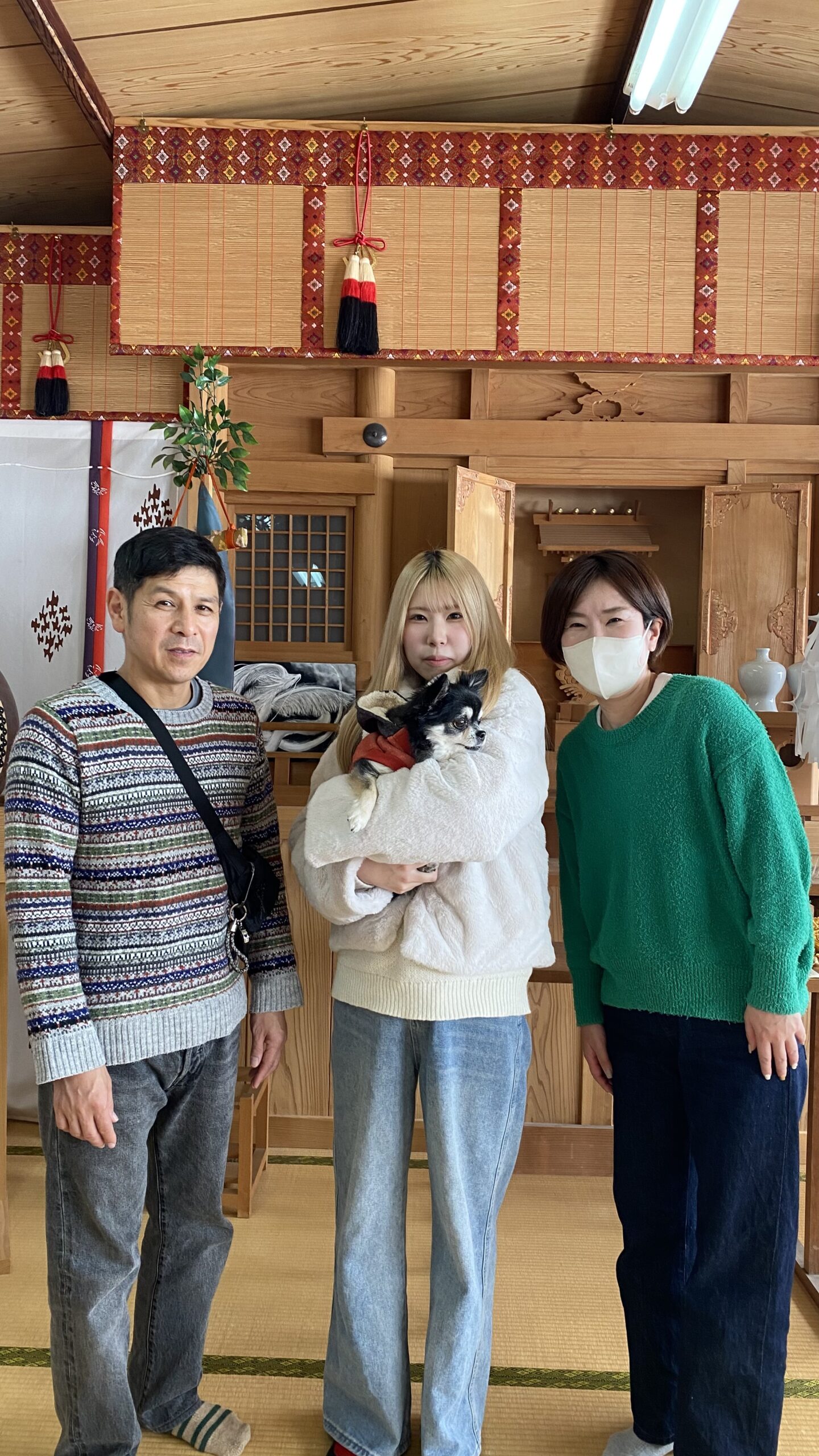 伝統的な和室の中で、手に小さな犬を抱えた白いセーターの女性が中央に立ち、両側にストライプのセーターを着た男性と緑のセーターを着たマスクをした女性が並んでいる様子。