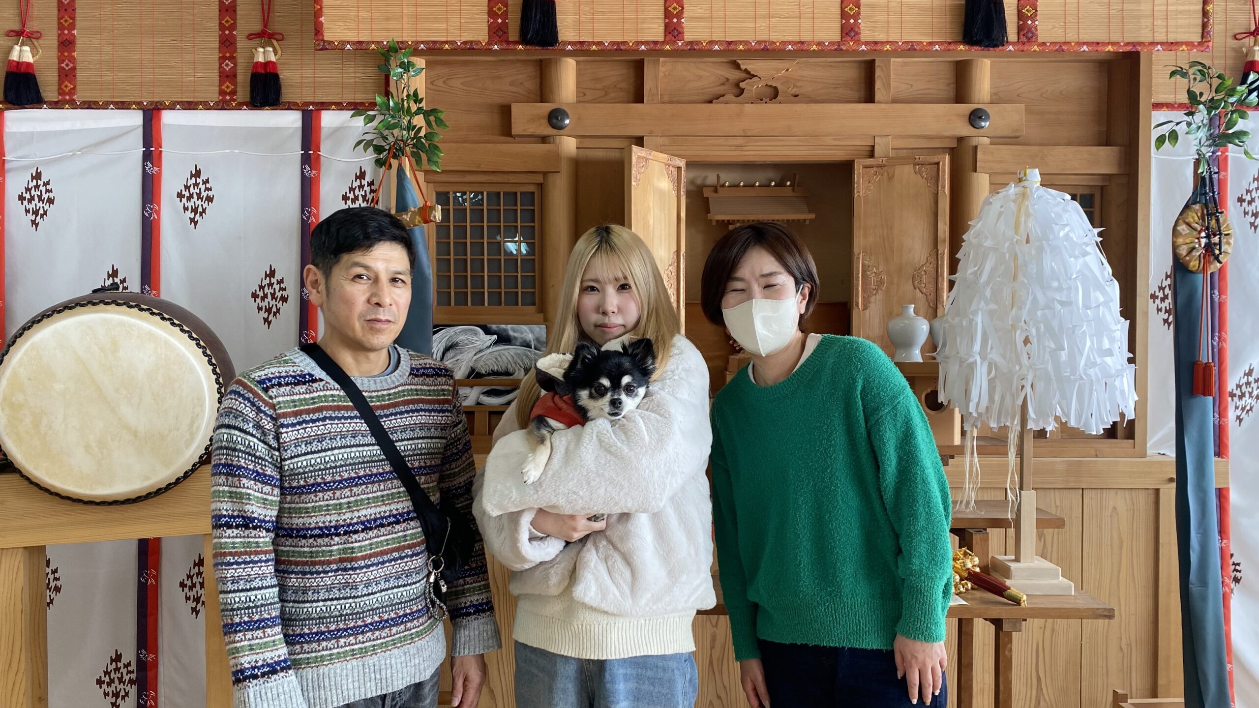 日本の伝統的な木造建築の室内で、緑のセーターを着たマスクの女性、白いふわふわのジャケットを着て小さな犬を抱える金髪の女性、模様の入ったセーターを着た男性の三人が並んで立っている様子。背景に大きな太鼓と装飾品が見える。