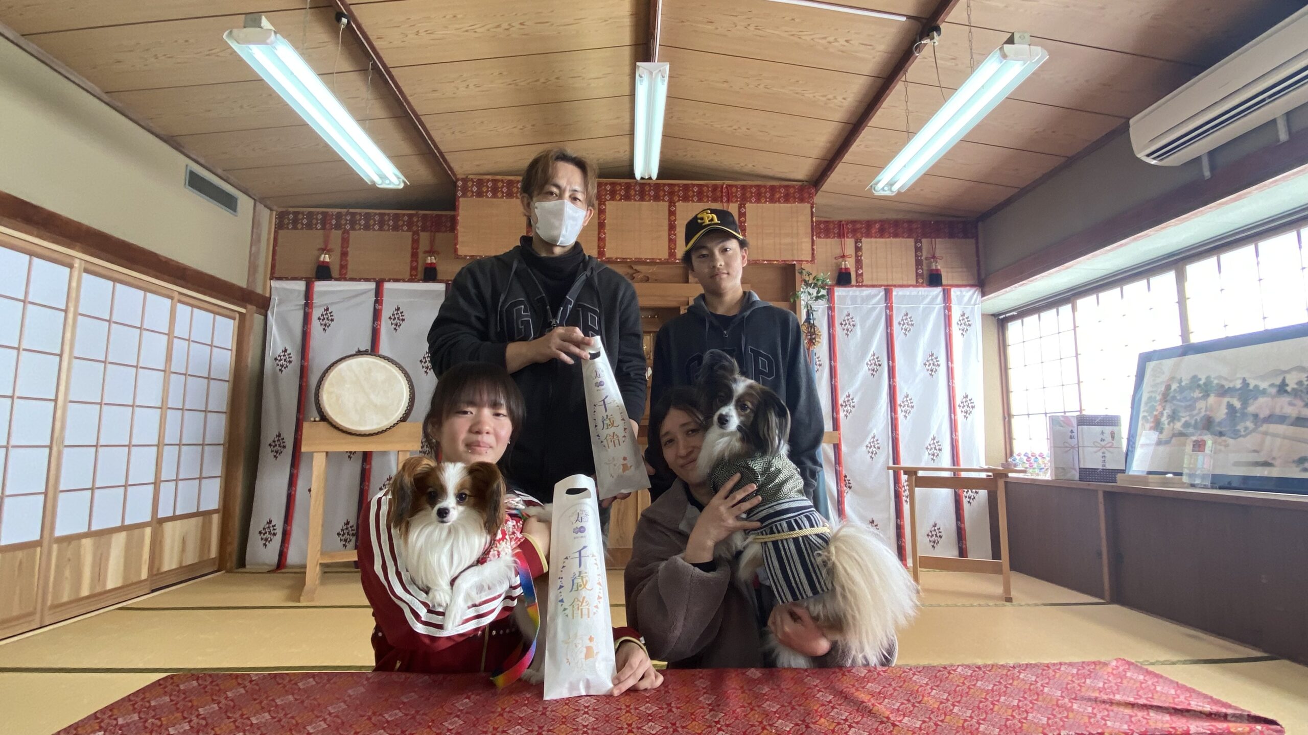 和風の部屋の中で4人の若者が二匹の小型犬を抱えて記念撮影している様子。背景に太鼓や掛け軸があり、畳が敷かれている。