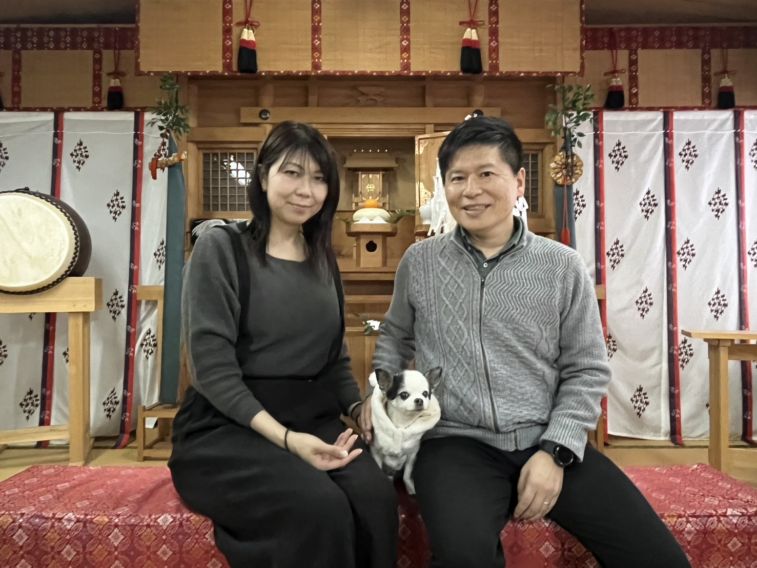神棚の前で座り、中央に小型犬を抱える男女が笑顔で並んでいる様子。